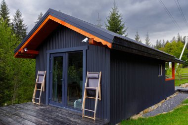 Bu gösterişli, modern kabin karanlık bir dış görünüme, büyük cam kapılara ve ahşap aksanlara sahiptir. Çevresindeki yeşillikle pürüzsüz bir şekilde harmanlanmış, sakin, doğal bir ortamda..