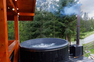 Bu resim, uzun ağaçların arasında yer alan ve sıcak ortam aydınlatmasıyla tamamlanmış doğal bir ortamda rahatlamayı sağlayan, suyla dolu sakin bir açık hava jakuzisi sergiliyor..