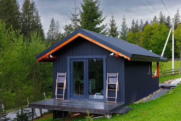 Canlı ağaçlar ve yeşilliklerin arasına yerleşmiş ahşap aksanlı şık siyah bir kulübe. Rahat bir açık hava oturuşu, doğanın sükuneti ve güzelliği arasında sakin anları davet eder..