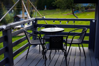 Yuvarlak cam tepeli, sicim ışıklarıyla süslenmiş metal sandalyeli bir masa içeren sakin bir açık hava güvertesi, yemyeşil yemyeşil ortamda huzur ve rahatlama anları..