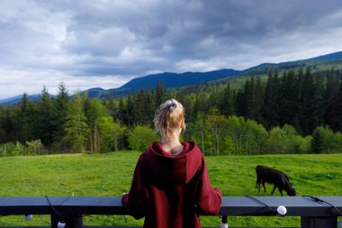 Bu sakin sahnede, kestane rengi kapüşonlu bir kadın yeşil dağların, sık ormanların ve otlayan bir ineğin resimsel manzarasına bakarak doğayla bir huzur ve bağlantı anı yaratıyor..