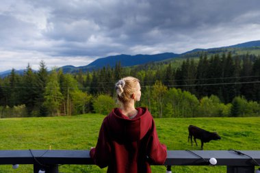 Görüntü, kahverengi kapüşonlu genç bir kadını, yemyeşil yemyeşil ve aşağıdaki tarlada otlayan huzurlu bir ineğin yer aldığı çarpıcı bir dağ manzarasına bakarken yakalıyor..