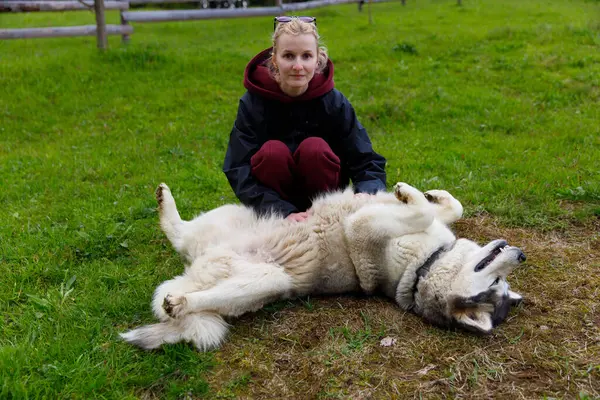 Resimli bir ortamda, bir kadın, yeşil bir alanda sırtüstü uzanan ve doğa arasındaki dostluk ve mutluluk bağını gösteren neşeli köpeğinin yanında oturur..