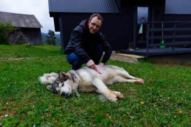 Bu sakin açık hava sahnesinde, bir adam köpeğiyle sevgi dolu bir şekilde iletişim kurar, yemyeşil bir arazi ve ahşap yapıların kırsal bir zemininde bağlarını gösterir..