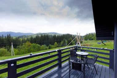 Sandalyeler ve bir masa ile süslenmiş büyüleyici bir balkon yuvarlanan tepeler ve bir orman manzarası sunuyor, açık hava toplantıları veya sessiz yansımalar için huzurlu bir ortam yaratıyor..