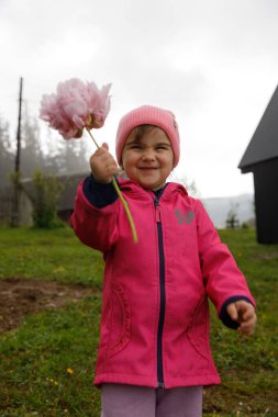 Pembe ceketli ve şapkalı mutlu bir genç kız, elinde pembe bir çiçek, masumiyeti ve çocukluk güzelliğini somutlaştıran açık hava ortamında neşeyle gülümsüyor..