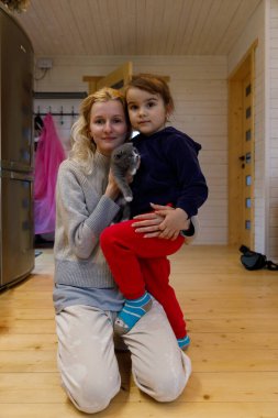 İçtenlikle dolu parlak bir evde, şefkatli bir kadının kucağında oturan genç bir kız, hem sevimli gri kedileriyle neşe saçıyor, güzel bir aile anı yakalıyor, sevgi ve birliktelik..