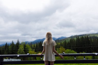 Huzurlu bir sahnede, zengin yeşillikler ve bulutlu gökyüzüyle çevrili balkondan yayılan dağlara bakan bir insan doğaya mükemmel bir kaçış sunuyor..
