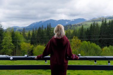 Görüntü, geniş dağlara bakan genç bir kadını ve balkondan yeşillik manzarasını yakalıyor. Doğa, huzur ve yansıma anlarının mükemmel bir karışımını sunuyor..