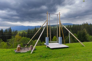 Bu büyüleyici görüntü, ahşap kirişler ve metal halkalarla inşa edilmiş, sıcacık bir ateş çukuruyla eşleştirilmiş bir salıncak setini gösteriyor..