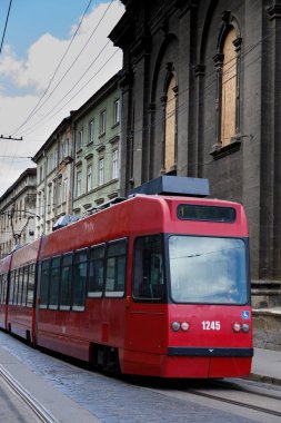 Görüntü, klasik mimariyle çevrili bir kaldırım taşı caddesinde duran çarpıcı bir kırmızı tramvayı yakalıyor. Sahne, tarihi cazibe ve modern teknolojinin ortasındaki kentsel geçişin evrimini yansıtıyor..