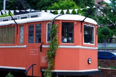 Bu resimde canlı turuncularla boyanmış, renkli bir tramvay, şenlikli kiraz kuşu ve yeşillikle süslenmiş, yemyeşil bir arkaplan ve kırsal taşlarla süslenmiş..