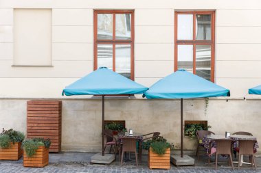 Görüntüde mavi şemsiyeli, davet masalı ve yemyeşil saksıları olan tuhaf bir açık hava kafe sergileniyor. Canlı bir şehir atmosferinde rahatlamak ve keyif almak için mükemmel bir ortam..