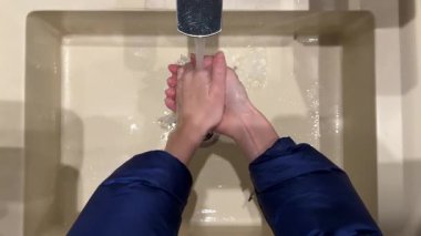 Üstten görüntülü, mavi ceketli kız ellerini açık sarı bir lavabo demirinden musluk içinde mikroplardan ve bakterilerden temizlenmiş temel hijyen sıvısı sabunu ile yıkıyor.