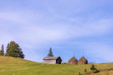 Bu pitoresk manzara, iki geleneksel saman yığınının yanında küçük ahşap bir kulübe sergiliyor. Nazikçe yuvarlanan tepeler ve yemyeşil ağaçlarla çevrili, sakin kırsal bir atmosfer yaratıyor..
