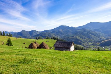 Bu güzel manzara yeşil tepeli, iki saman yığını, geleneksel bir kulübe ve güneşli bir günde geniş dağ manzaralı kırsal güzelliğin özünü yakalar..