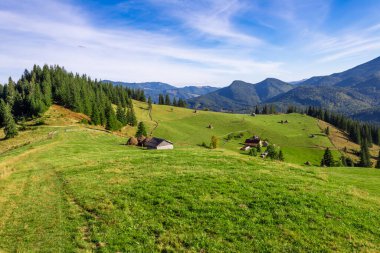 Göz kamaştırıcı bir panorama yeşil alanlar, kır evleri ve yüksek ağaçlar, uzak dağlar parlak mavi gökyüzünün altında sakin bir zemin oluşturur..