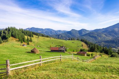 Geleneksel saman yığınları, kırsal ahşap ahırlar ve yüksek çam ağaçları ile süslenmiş yeşil yuvarlanan tepeleri gösteren sakin bir manzara, hepsi berrak bir gökyüzünün altındaki görkemli dağ sıraları tarafından çerçevelenmiş..
