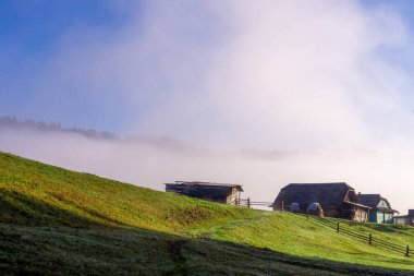 Bereketli yeşillikler ve berrak mavi gökyüzü ile çevrili yuvarlanan tepelerde yuva yapmış kırsal çiftlikleri kaplayan sisi tasvir eden huzurlu bir sabah manzarası sükunet ve güzelliği çağrıştırıyor..