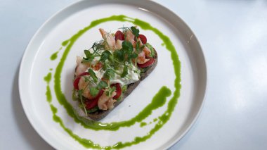 Bu nefis yemek, üzerinde taze karides, canlı vişne domatesleri, kremalı avokado ve yeşil sosla zarif bir şekilde sunulmuş yeşil bir ekmek tabanı içerir..