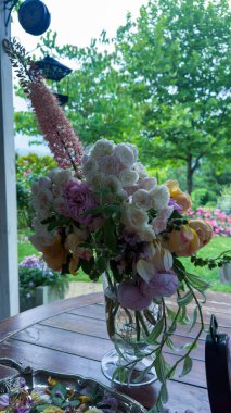 Bu güzel çiçek aranjmanı cam bir vazoda renkli bir çiçek dizisi sergiliyor, yeşillik ve çiçeklerle dolu güzel bir bahçe zeminine kurulmuş..