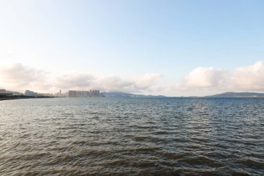 Sakin suların manzarası bulutlu bir gökyüzünü yansıtıyor, sahil boyunca uzanan kentsel binalar, doğanın uyumlu bir karışımını yaratıyor güzellik ve insan mimarisi mükemmel bir bütünlük içinde..