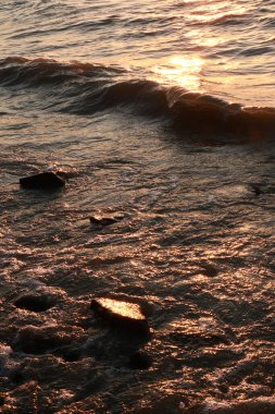 Görüntü, batan güneş tarafından aydınlatılan dalgaların ritmik dansını güzel bir şekilde sergiliyor. Kıyı şeridine yerleştirilmiş pürüzsüz kayalarla su üzerinde altın renkleri yansıtıyor..