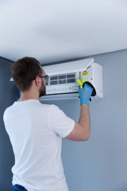 Görüntü, duvardaki klimayı temizlemek için bez kullanan birini gösteriyor. Bu temizlik süreci iyi hava kalitesi ve optimal performansı korumak için gereklidir..