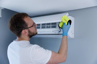 Odaklanmış bir birey sarı bir bez kullanarak havalandırma ünitesini dikkatlice temizliyor ve rahat bir iklim için düzenli bakımın önemini vurguluyor..