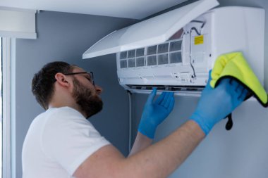 Bu resimde, gözlüklü ve sakallı bir adam evin havalandırmasını dikkatlice temizlerken aynı zamanda tek kullanımlık eldiven takıyor..