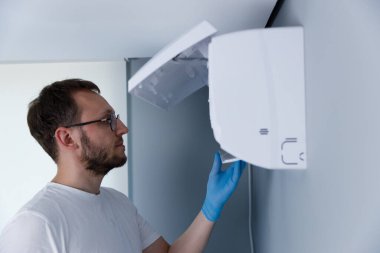 Görüntü, duvarlara monte edilmiş bir cihazı inceleyen, temiz bir ortamda modern bakım uygulamaları ve güvenlik önlemlerini vurgulayan, günlük beyaz gömlekli bir teknisyeni yakalıyor..