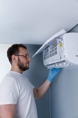 Mavi eldiven giyen bir teknisyen, evlerdeki en uygun işlevsellik ve hava kalitesi için düzenli bakımın önemini vurgulayarak, kapalı bir havalandırma ünitesini denetliyor..