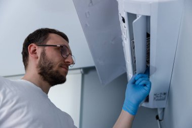 Görüntü, beyaz gömlekli ve mavi eldivenli çalışkan bir kişinin düzgün çalışma ve temizliği sağlamak için bir havalandırma sistemini dikkatlice inceleyip sürdürdüğünü gösteriyor..