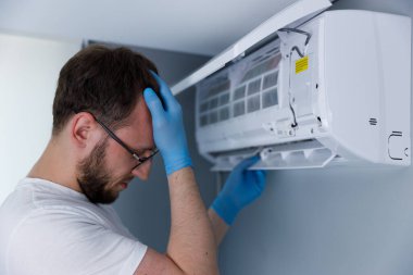 Klima tamir teknisyeni, arka planı hafif olan çağdaş bir ortamda stres ve sorun çözme sorunlarını temsil ederek, klima ünitesini tamir etmekte güçlük çekiyor.