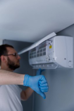 Görüntü mavi eldivenli bir teknisyeni gösteriyor. Klima ünitesinden memnuniyetsizliğini başparmağını kaldırarak ifade ediyor. Ayarlama bir hayal kırıklığı hissi veriyor..