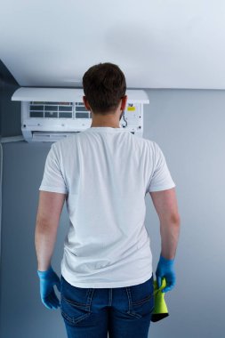 Görüntü, gri bir duvara monte edilmiş klima ünitesini muhafaza etmeye hazır, mavi eldivenler ve düz beyaz bir tişört giyen, temiz kesimli bir saç stiline sahip bir kişiyi gösteriyor..