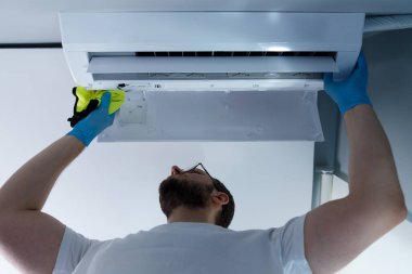 Mavi eldivenli odaklanmış bir adam havalandırma ünitesini temizliyor. Parlak bir bez kullanarak, cihazın etkinliğini ve sağlık yararları için daha iyi kapalı hava kalitesini sağlamak için dikkatle muhafaza eder..