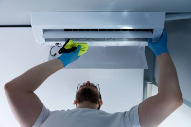Görüntü, klima ünitesini özenle koruyan bir adamın mikro fiber bez kullanırken koruyucu eldiven giydiğini ve kapalı hava kalitesinin önemini vurguladığını gösteriyor..