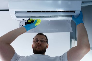 Mavi eldiven giyen odaklanmış bir birey, yukarıdaki havalandırma ünitesini parlak sarı bir bezle temizliyor ve düzenli bakımın konfor ve temizlik açısından önemini vurguluyor..