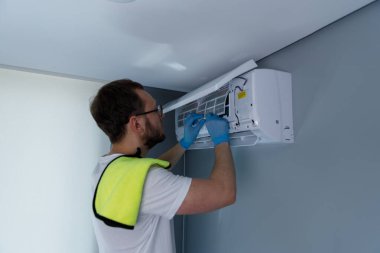 Becerikli bir teknisyenin duvara monte edilmiş bir havalandırma ünitesinde çalışırken görülmesi, rutin bakımların etkin soğutma ve kapalı hava kalitesi açısından önemini vurguluyor..