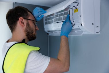 Görüntü, klima sistemini tamir etmeye odaklanmış bir teknisyeni yakalıyor. Mavi eldiven ve parlak sarı bir yelek giyiyor. Birim bileşenleri üzerinde çalışırken güvenliği vurguluyor..