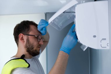 Mavi lastik eldiven giyen bir adam, bir alet sorununu çözerken derin düşüncelere dalmış gibi görünür. Cihazı kullanırken hayal kırıklığına uğramış görünüyor. Bu da teknik bir arıza olduğunu gösteriyor..