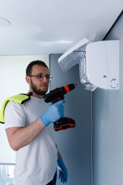 Odaklanmış bir birey, iyi ışıklandırılmış, modern bir odada duvara bir klima yerleştirirken ve görev için eldiven ve giysi giyerken bir elektrikli matkap kullanıyor..