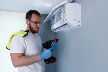 Görüntü, elinde matkapla odaklanmış bir adamın omuzunda eldiven ve havlu varken klima bakımına hazırlandığını gösteriyor. Bu da temiz bir ortamda profesyonel hizmet olduğunu gösteriyor..