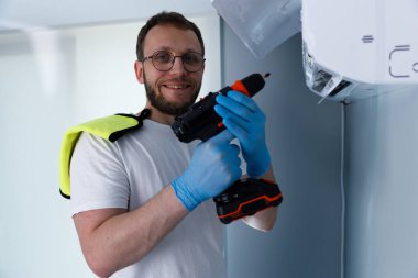 Elektrikli matkapla donatılmış ve eldiven giyen gülümseyen bir teknisyen klima bakımına hazır bekliyor. Parlak ve temiz bir ortam profesyonelliği ve hazırlığı vurguluyor.
