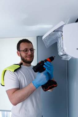 Görüntü, eldiven ve matkapla donatılmış bir teknisyenin, bir havalandırma ünitesinin bakımını üstlenmesi, ev iyileştirme görevlerinde çalışkanlığı ve uzmanlığı simgeliyor..