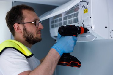 Kendini işine adamış bir teknisyen, bir klima ünitesine elektrikli matkap kullanarak özenle bakım yapıyor. Çağdaş bir iç mekanda detaylara titizlikle dikkat çekiyor..