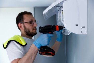 Bu resimde, sakallı odaklanmış bir teknisyen duvardaki beyaz klimayı tamir etmek için bir matkap kullanıyor, eldivenlerle donatılmış ve korunmak için bir güvenlik bezi kullanıyor..