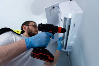 Görüntü, koruyucu eldiven giyen bir teknisyenin beyaz bir duvara monte edilmiş bir klima üzerinde çalışmak için matkap kullanmasını ve tamiratın hassasiyetini vurguluyor..