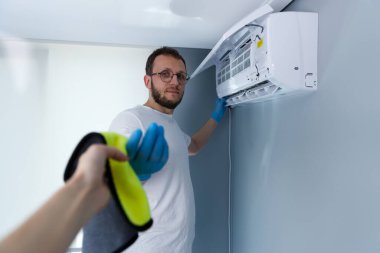 Görüntü, temiz bir bez tutarken eldiven giymiş bir adamı bir havalandırma ünitesine girerken yakalıyor. Bu da düzenli bakımın ideal performans ve hijyen için önemini gösteriyor..